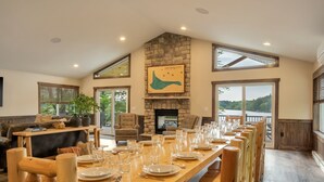 A dining area with a long wooden table set for a meal, a stone fireplace, and large windows offering a view of the outdoors. - Lake Front Livin by PMR, 8BR Lakefront, Private Beach & Dock, Sleeps 26 (Lake Harmony)