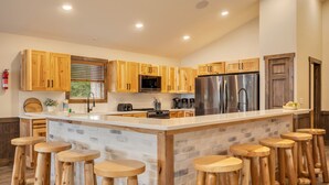 A modern kitchen with wooden cabinets, a central island with stools, stainless steel appliances, and a window with blinds. - Lake Front Livin by PMR, 8BR Lakefront, Private Beach & Dock, Sleeps 26 (Lake Harmony)
