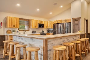 A modern kitchen with wooden cabinets, a central island with stools, stainless steel appliances, and a window with blinds.