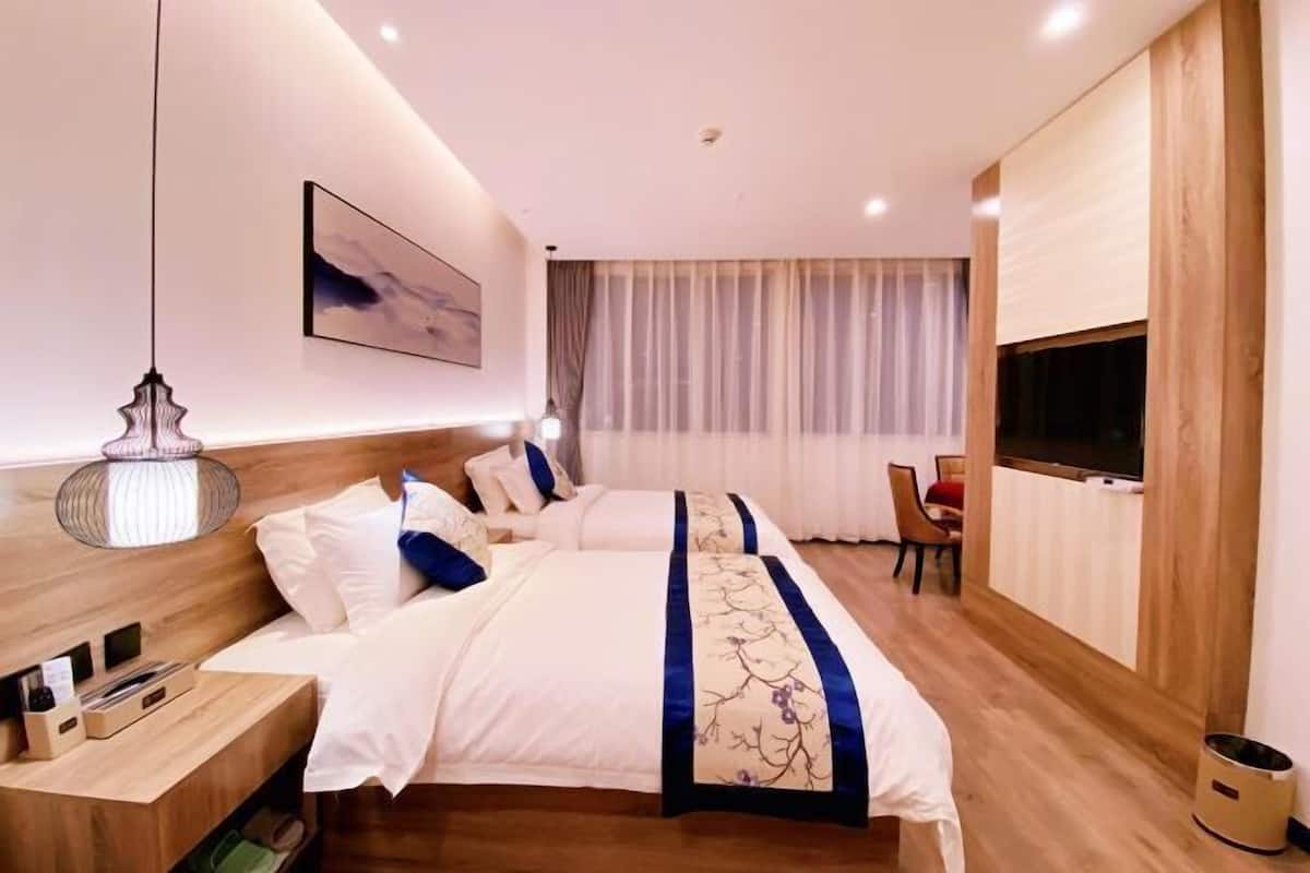 A hotel room with a large bed, a bedside table with a lamp, a TV mounted on the wall, and a window with curtains.