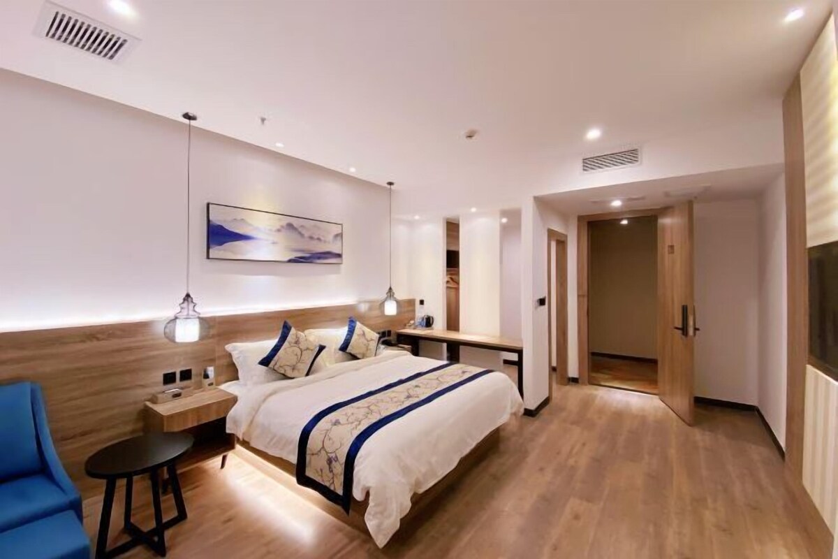 A modern hotel room with a wooden floor, a bed with a blue and white bedspread, a bedside table with a lamp, and a painting on the wall.