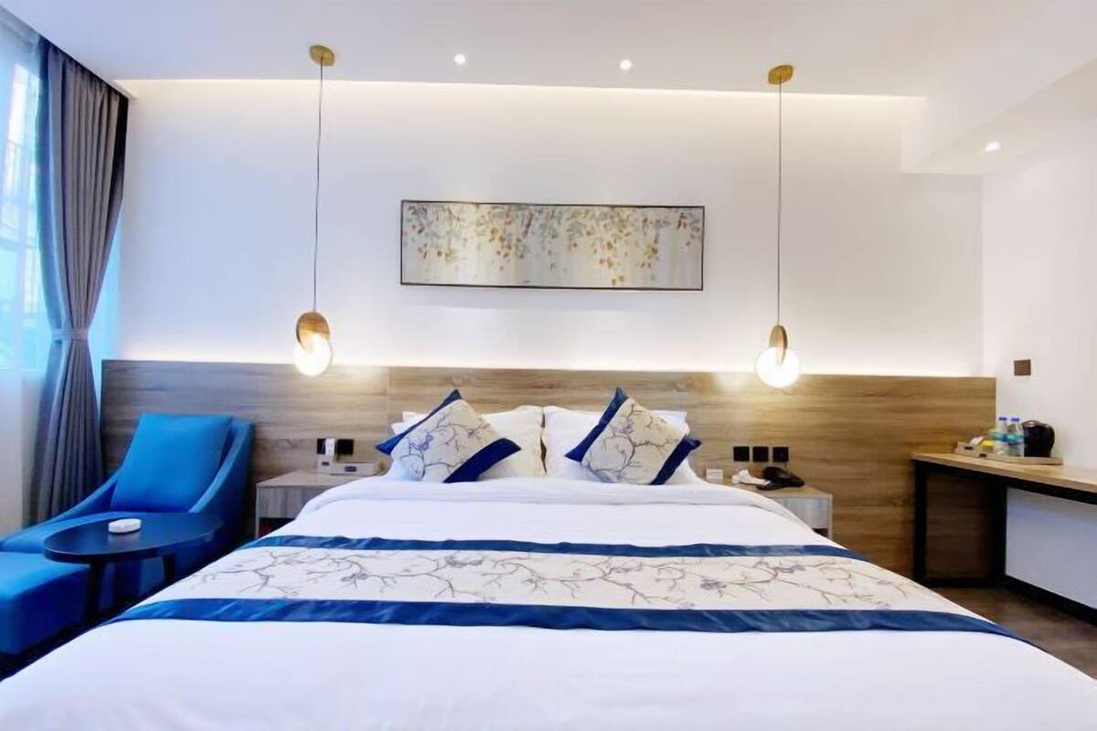 A modern hotel room with a large bed, a bedside table, a desk, a chair, and a hanging light fixture.