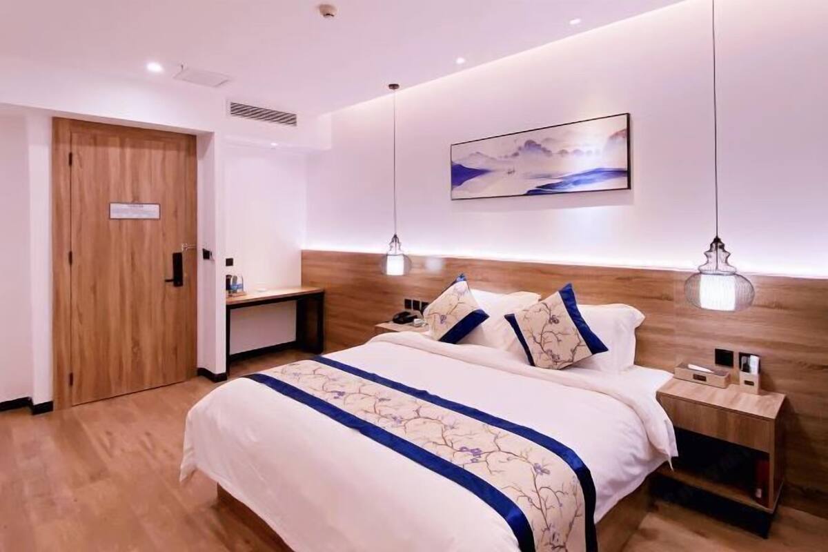 A modern hotel room with a wooden headboard, a large bed with a floral bedspread, bedside tables, and a wall-mounted artwork.