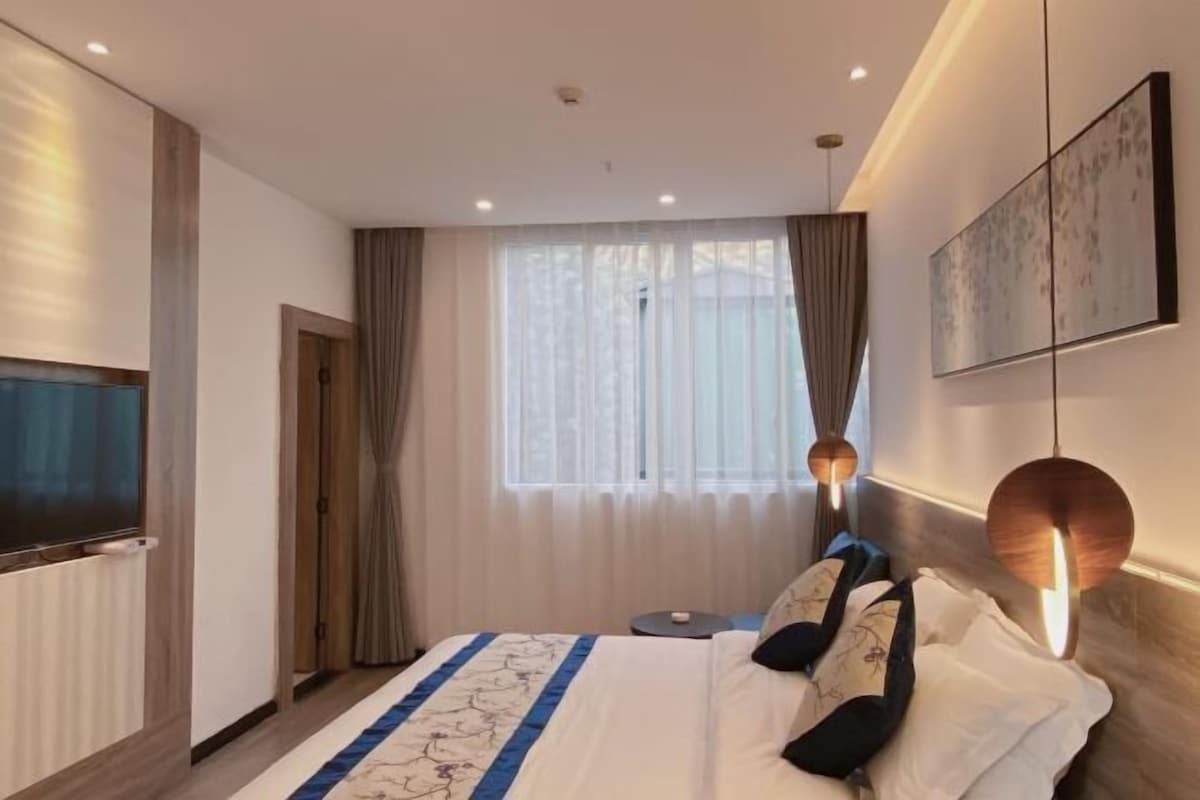 A hotel room with a bed, a TV, a window with curtains, and a wall-mounted artwork.