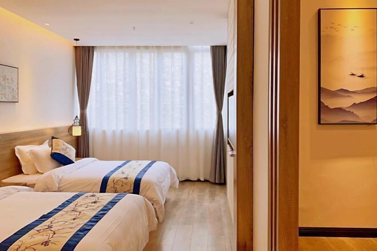 A hotel room with two beds, a large window with curtains, a painting on the wall, and a wooden floor.
