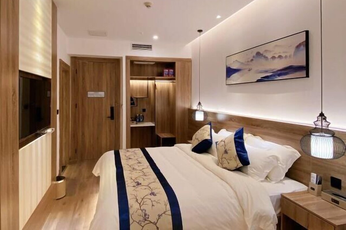 A modern hotel room with a large bed, a wooden wardrobe, a small desk, and a wall-mounted TV.