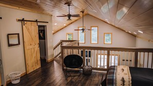 Une chambre spacieuse de style loft, avec un plafond haut, des poutres apparentes, un balcon avec une balustrade, un grand lit, une chaise et un ventilateur de plafond.