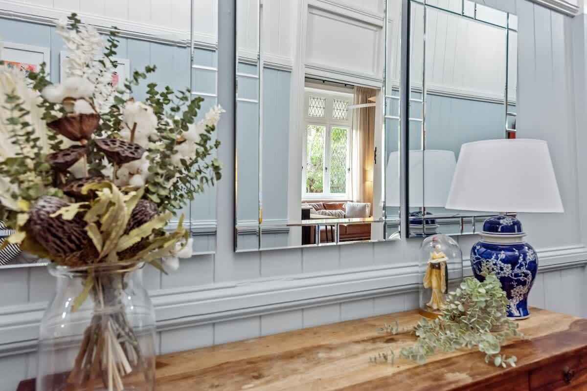 A room with a wooden dresser, a vase with dried flowers, a blue vase, and a mirror reflecting a window with a view of a room.
