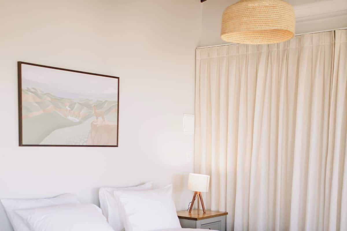 A neatly made bed with white linens in a room with a wooden ceiling and a large window with curtains.
