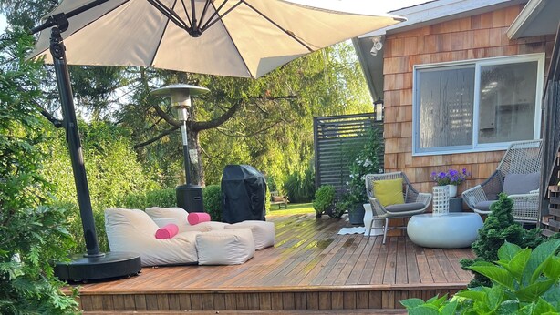 Een houten terras met tuinmeubilair, een pergola met een parasol en een tuin met staptegels.
