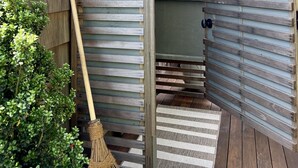 Een buitendouche van hout met een hangmand vol bloemen, een bezem en een gestreept kleed.