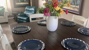 Ein Esstisch ist mit blau-weiß karierten Tellern gedeckt, umgeben von weißen Stühlen. In der Mitte steht eine weiße Vase mit bunten Blumen.