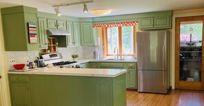 A kitchen with green cabinets, a stainless steel refrigerator, a stove, and a window with a red and white valance.