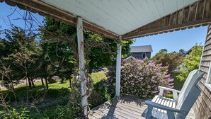 A wooden deck with a white chair, surrounded by greenery and a house in the background.