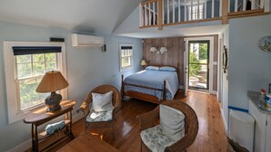 A bedroom with a loft bed, a wooden chair, a bedside table with a lamp, and a window with a view of greenery.