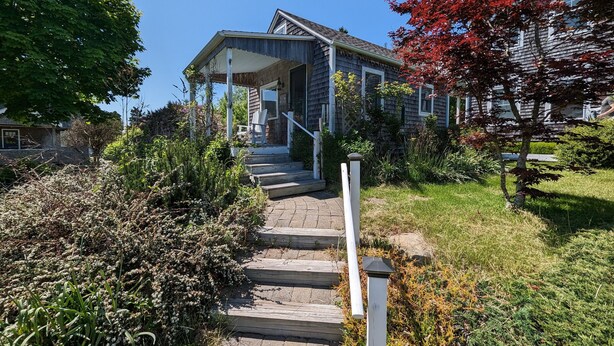 A house with a covered porch, steps leading to the entrance, and a garden with various plants and a small tree with red leaves.