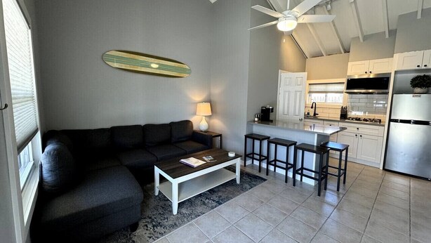 A modern living room with a dark sofa, a coffee table, and a kitchen area with a bar stool and a refrigerator. - Brand New Beach Condo 2BD 1BA w/Parking (Point Pleasant Beach)