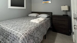 A bedroom with a bed, a nightstand, a wall-mounted air conditioner, a window with blinds, and a colorful artwork. - Brand New Beach Condo 2BD 1BA w/Parking (Point Pleasant Beach)