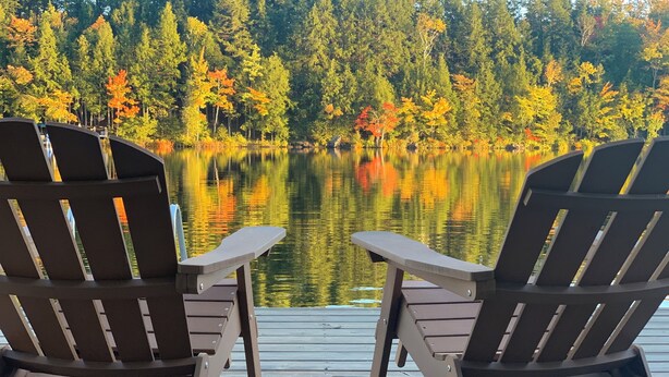 Deux fauteuils Adirondack sont placés face à un lac paisible, avec le feuillage automnal en arrière-plan.