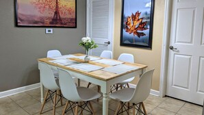Un comedor con mesa de madera, sillas blancas, un jarrón con flores y cuadros en las paredes.