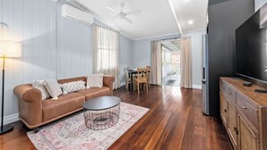 Un salon avec un canapé en cuir marron, une table basse ronde, un téléviseur à écran plat, un meuble en bois et un coin repas comprenant une table et des chaises.