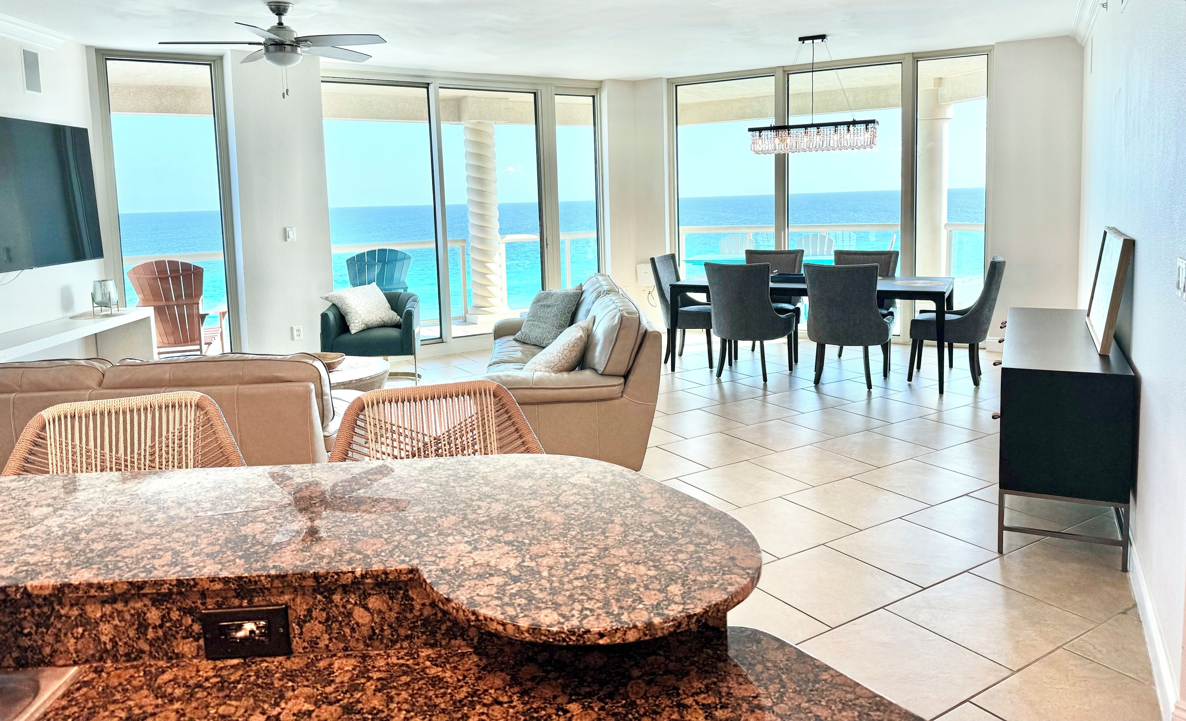 A spacious living room with a large window overlooking the ocean, a ceiling fan, and a dining area with chairs and a table.