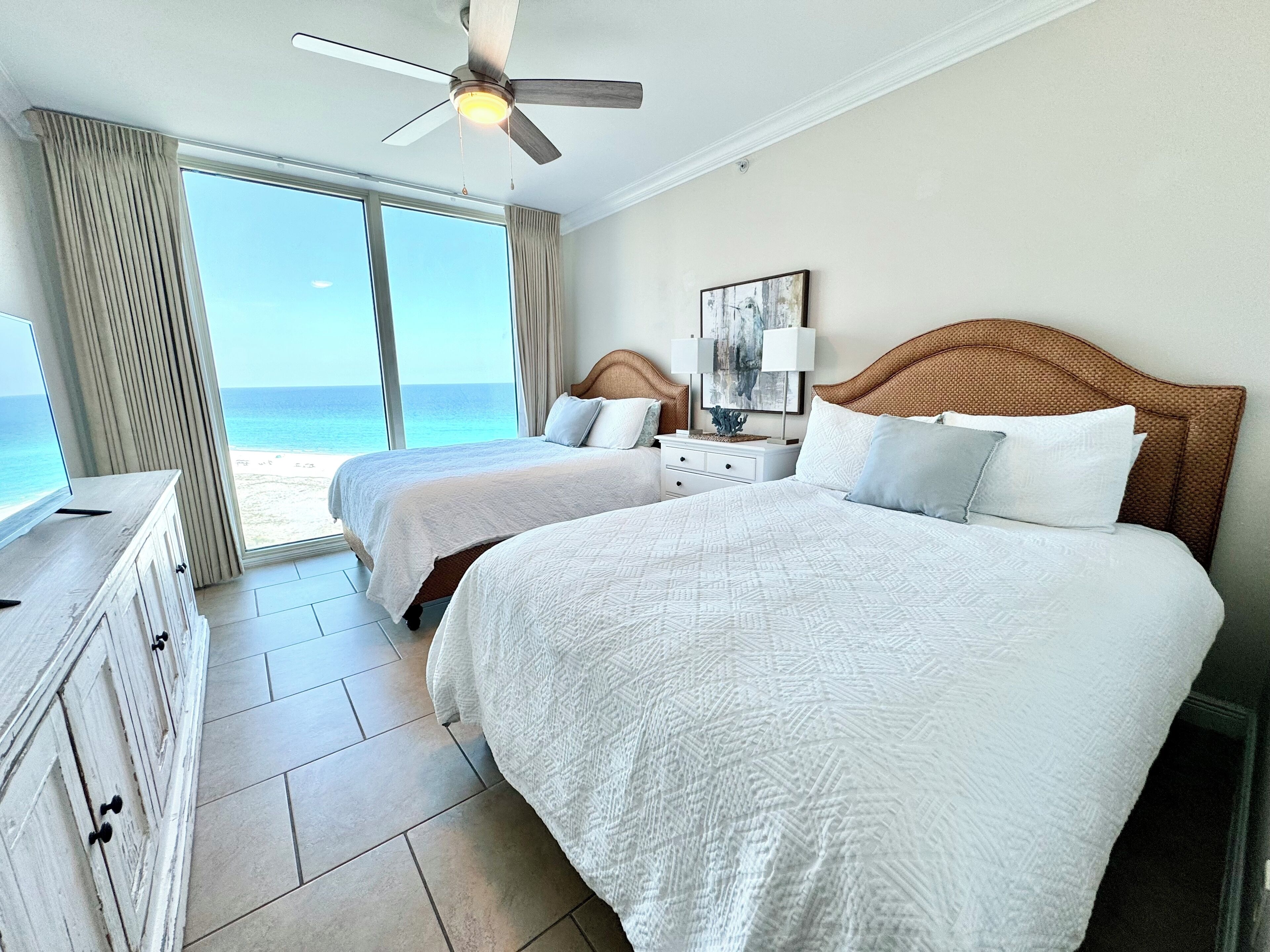 Une chambre d’hôtel avec deux lits, un ventilateur de plafond, une table de chevet avec une lampe et une vue sur la plage.
