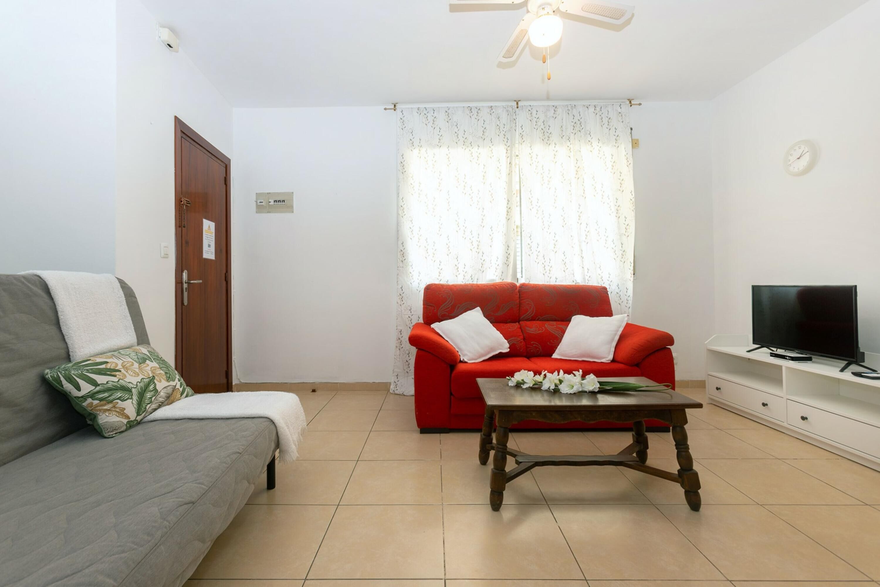 A living room with a grey sofa, a red sofa, a wooden coffee table, a television on a white stand, and a ceiling fan.