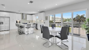 Una cocina moderna con armarios blancos, una isla central con taburetes de bar y grandes ventanales con vistas a palmeras y edificios.