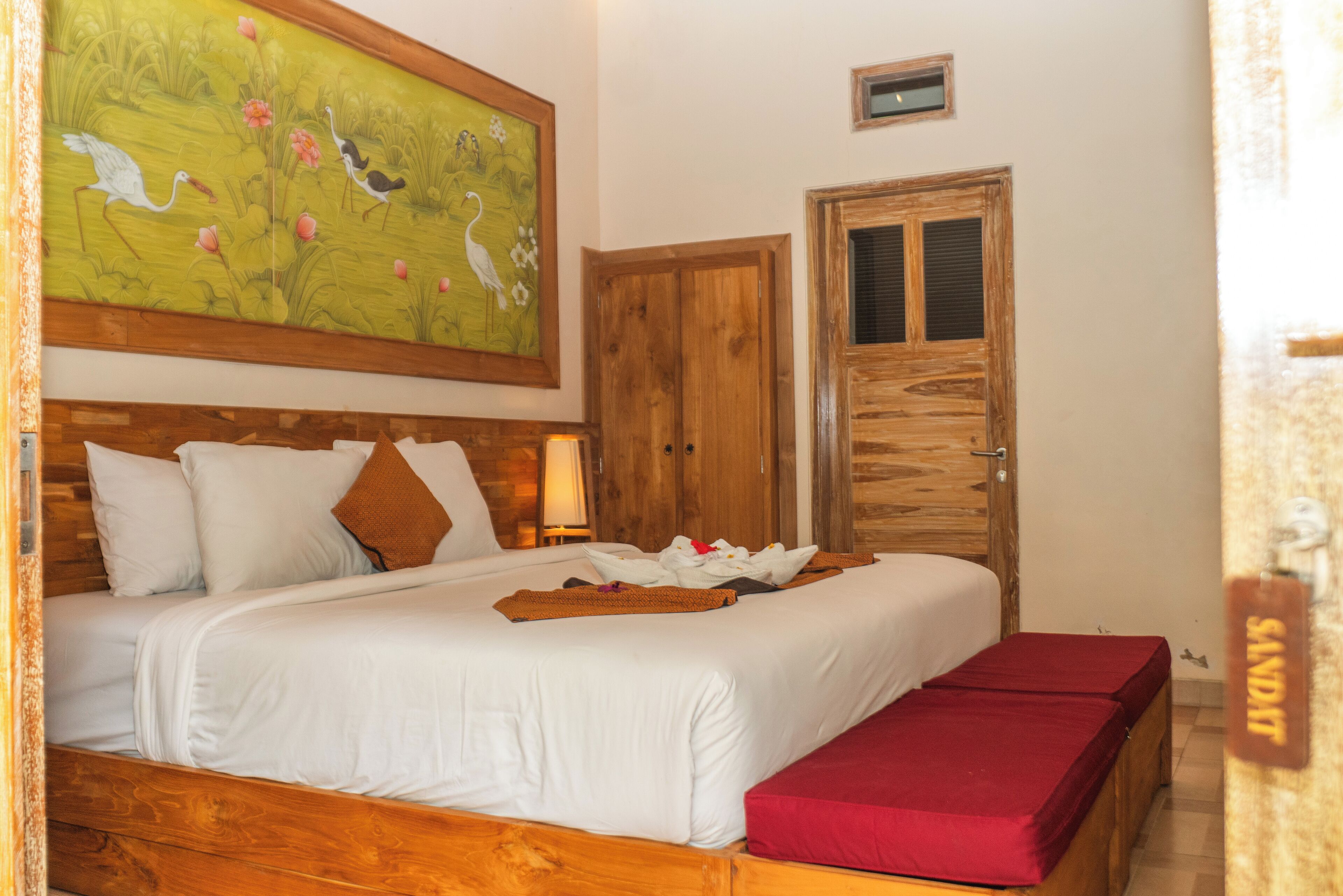 A neatly made bed with white linens and a red bench, a wooden door, and a painting of birds on the wall.