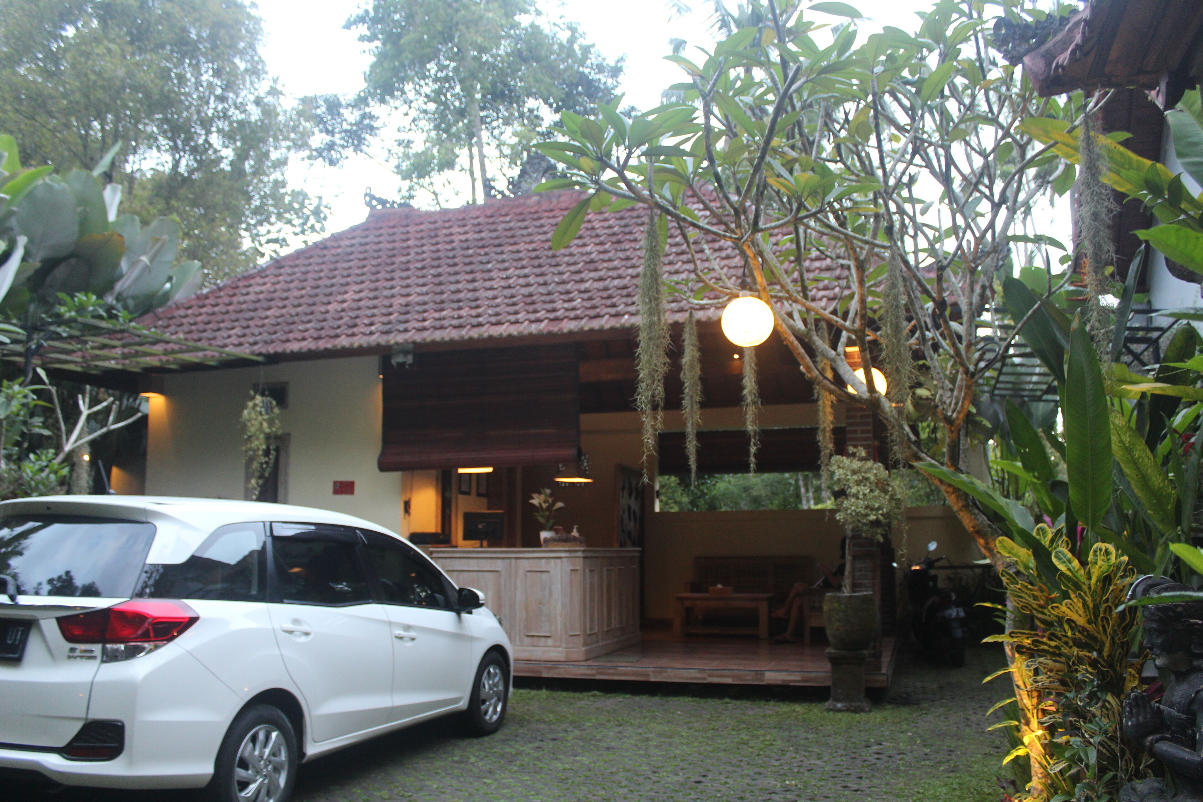 A white car parked in front of a building with a red-tiled roof, a porch, and a garden with hanging plants.
