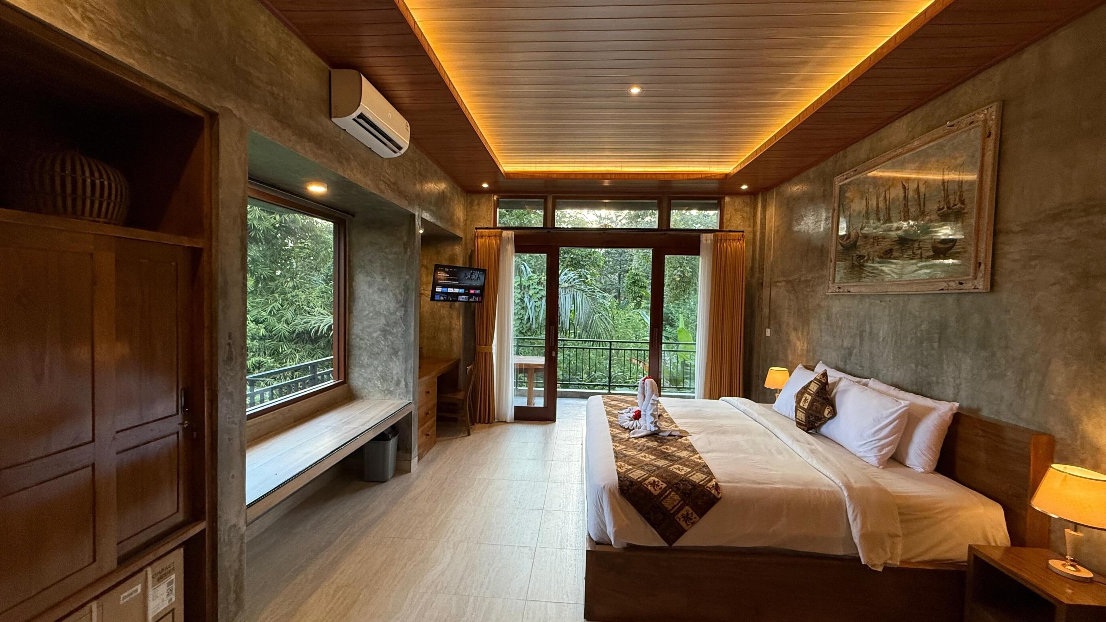 A bedroom with a large bed, a TV, a bench, and a balcony with a view of greenery.