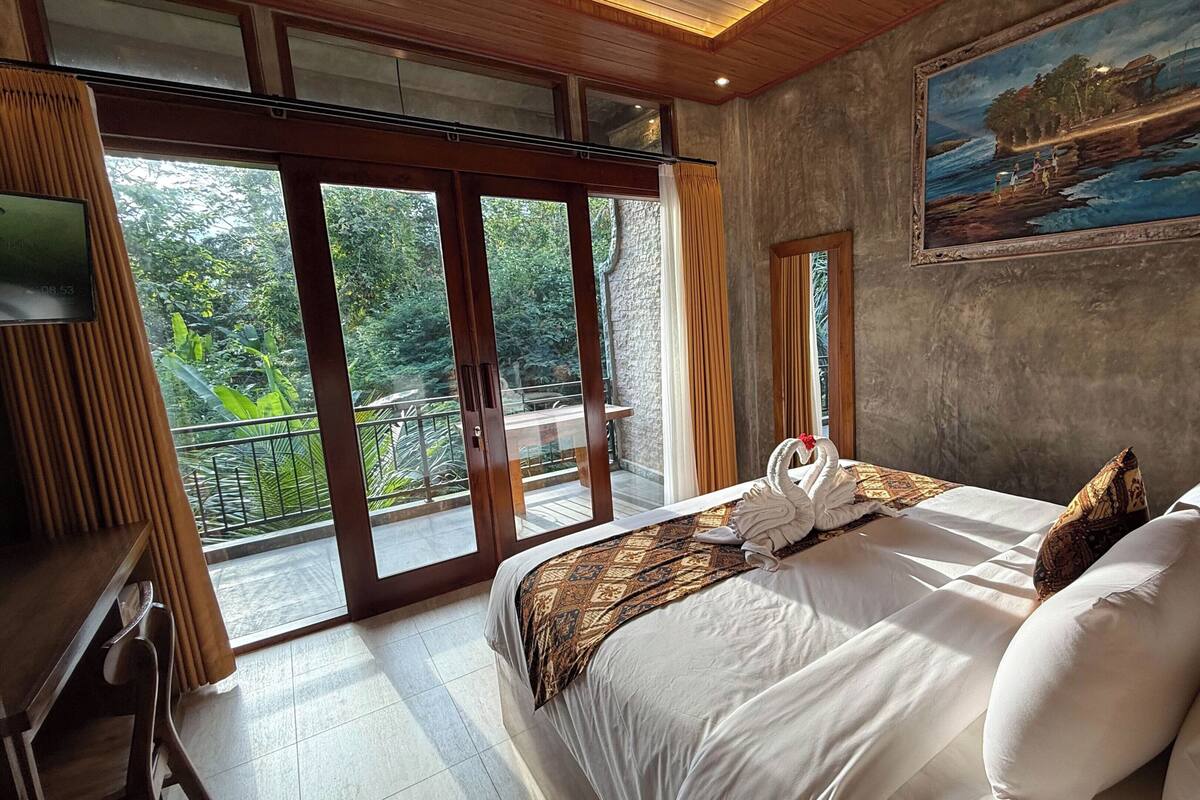 A hotel room with a bed, a painting, a balcony with a view of greenery, and a television.