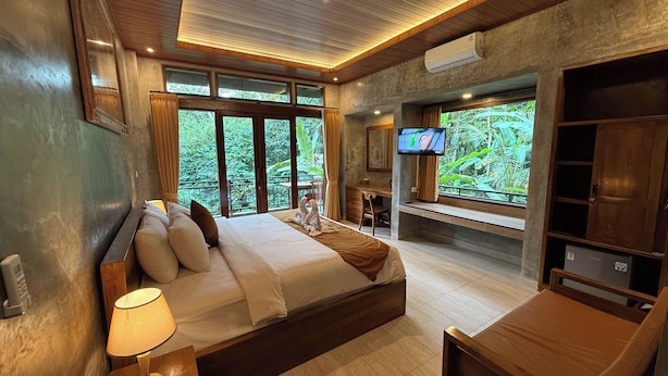 Un dormitorio con cama, escritorio, televisor y balcón con vistas a la vegetación.