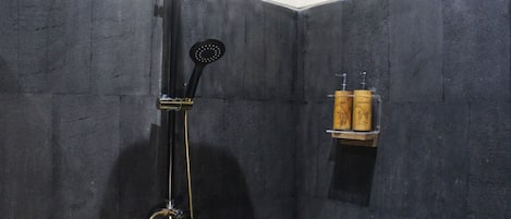 A shower with a handheld showerhead and a wall-mounted faucet. There is a shelf with two wooden bottles.