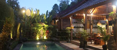 A pool area with a wooden deck, surrounded by tropical plants and a covered seating area with tables and chairs.