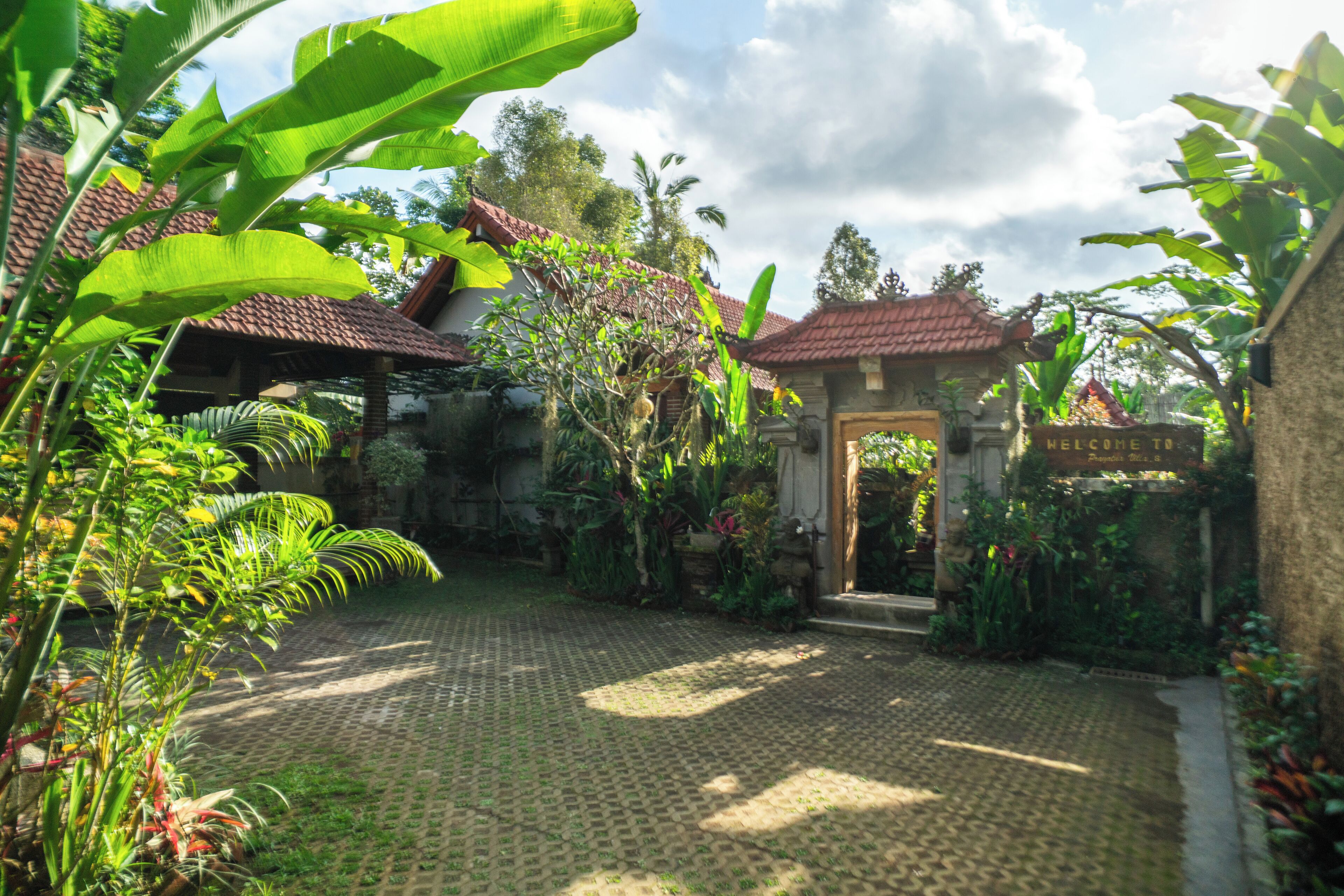 A courtyard with a paved walkway, a red-tiled roof structure, and lush tropical vegetation.