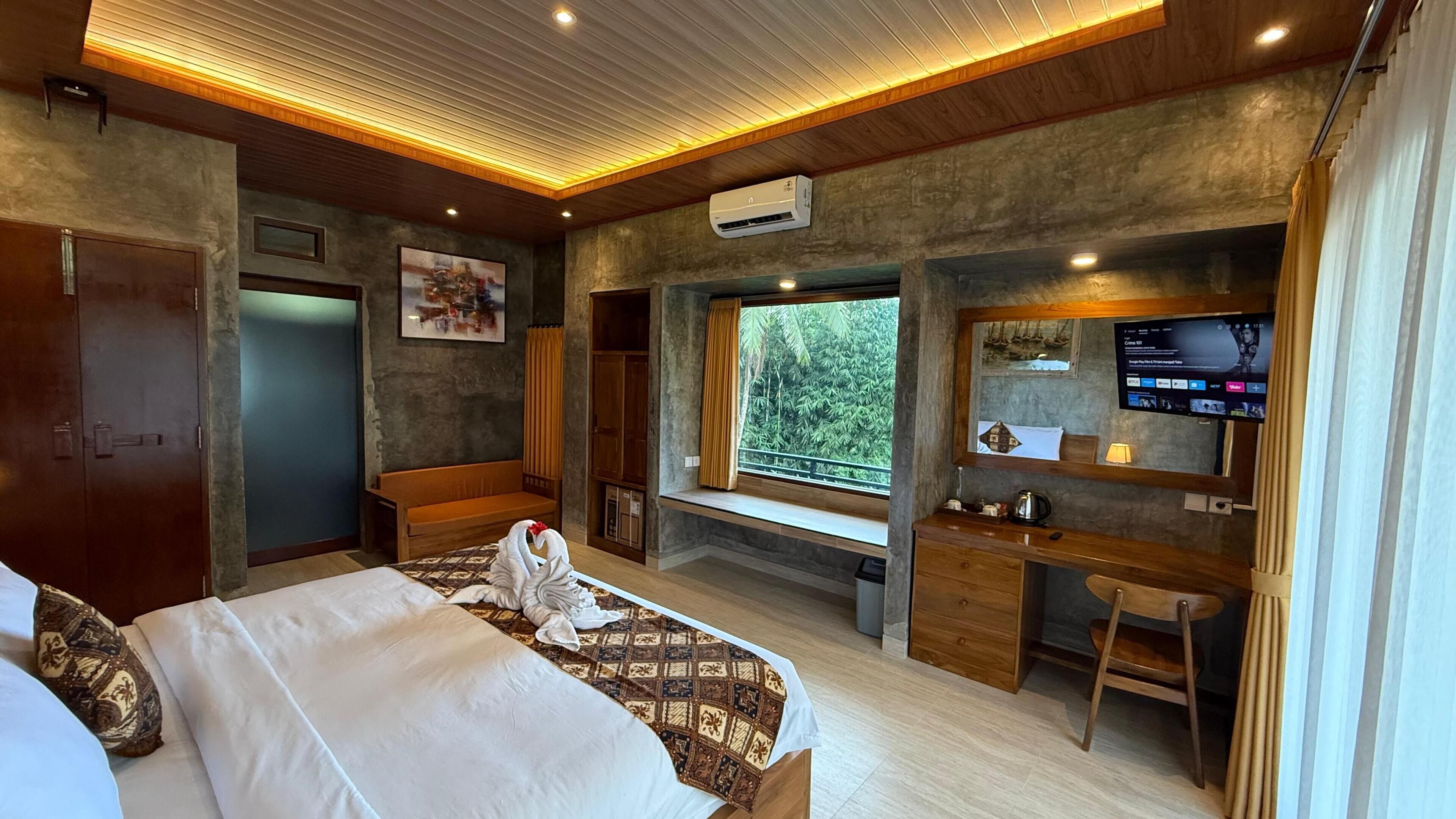 A hotel room with a bed, a desk, a TV, and a window with a view of greenery.