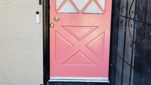 Una puerta rosa con un ventanal de diseño de diamantes, un cartel con el número 2, un felpudo con un mensaje de bienvenida y una celosía de seguridad negra a la derecha.