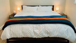 Una cama bien hecha con edredón blanco y manta a rayas de colores, dos lámparas de noche y una pared decorada con motivos de lunas.