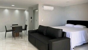 A modern hotel room with a black leather sofa, a single bed with white linens, a small dining table with chairs, and an air conditioning unit mounted on the wall. - Atlântico Vista Mar Deluxe (Fortaleza)