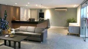A modern living room with a sofa, a coffee table, and a Christmas tree. There is a reception desk with a TV and a clock on the wall. - Atlântico Vista Mar Deluxe (Fortaleza)