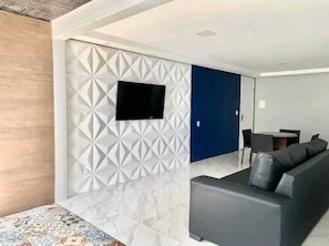 A modern living room with a flat-screen TV mounted on a geometric-patterned wall, a dark-colored sofa, and a small table with chairs. - Atlântico Vista Mar Deluxe (Fortaleza)