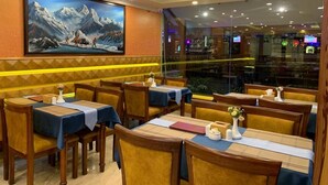 Un restaurante con mesas y sillas de madera, un cuadro grande de un paisaje montañoso en la pared y vistas de la ciudad a través de la ventana.