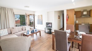 Una sala moderna con un sofá, una mesa de centro y una mesa de comedor preparada para cuatro comensales. Un televisor está montado en la pared, y se ve una cocina al fondo.