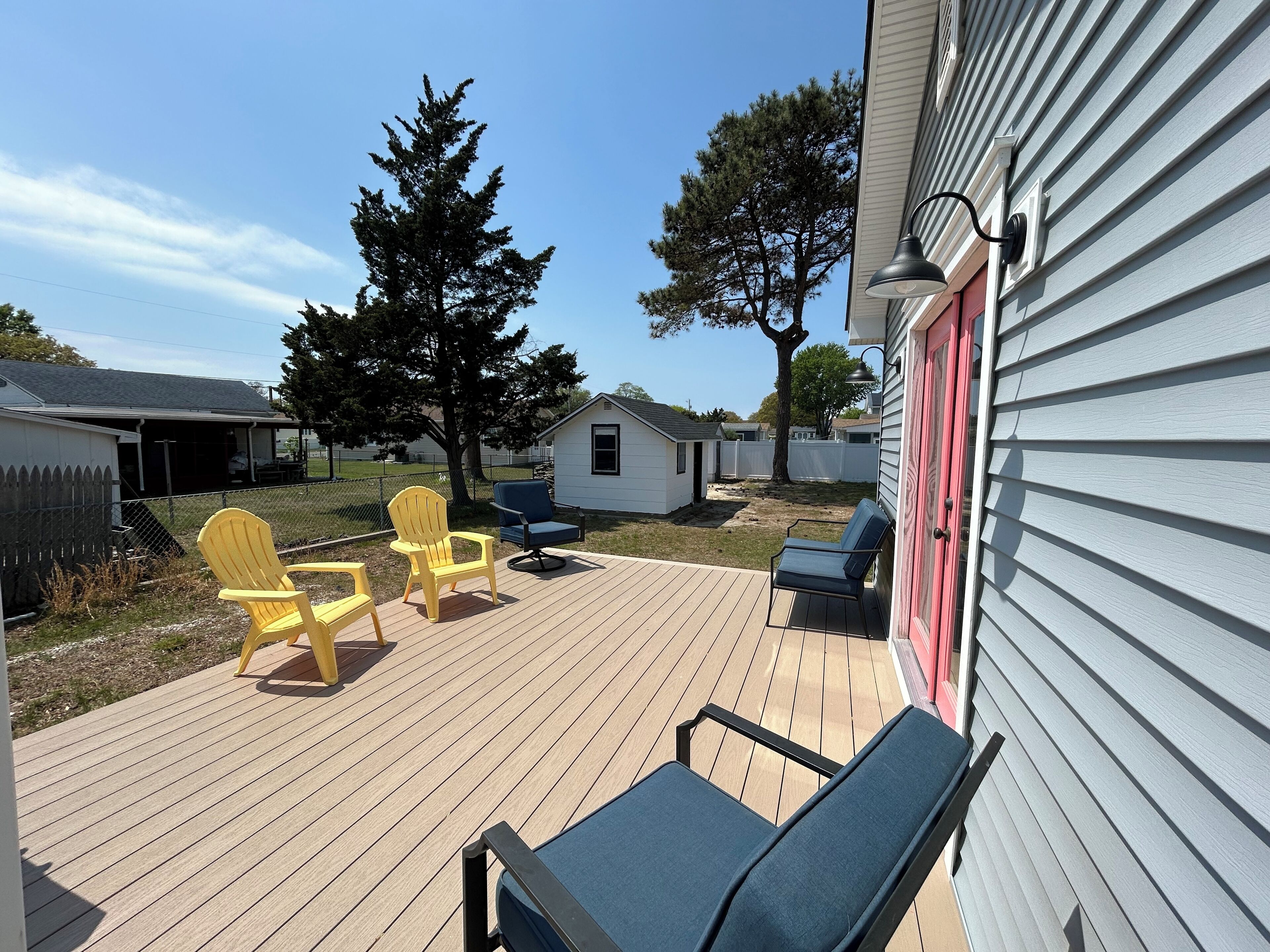 A deck with outdoor seating, including two yellow Adirondack chairs and one blue lounge chair, surrounded by a wooden deck and a white building with a red door.
