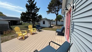 Um deck com área de estar ao ar livre, incluindo duas cadeiras Adirondack amarelas e uma poltrona azul, cercado por um deck de madeira e um edifício branco com porta vermelha.