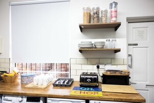 Una cocina con encimera de madera, un tostador, una tabla de cortar y diversos utensilios de cocina. Hay estantes sobre la encimera con frascos y una lata de avena.