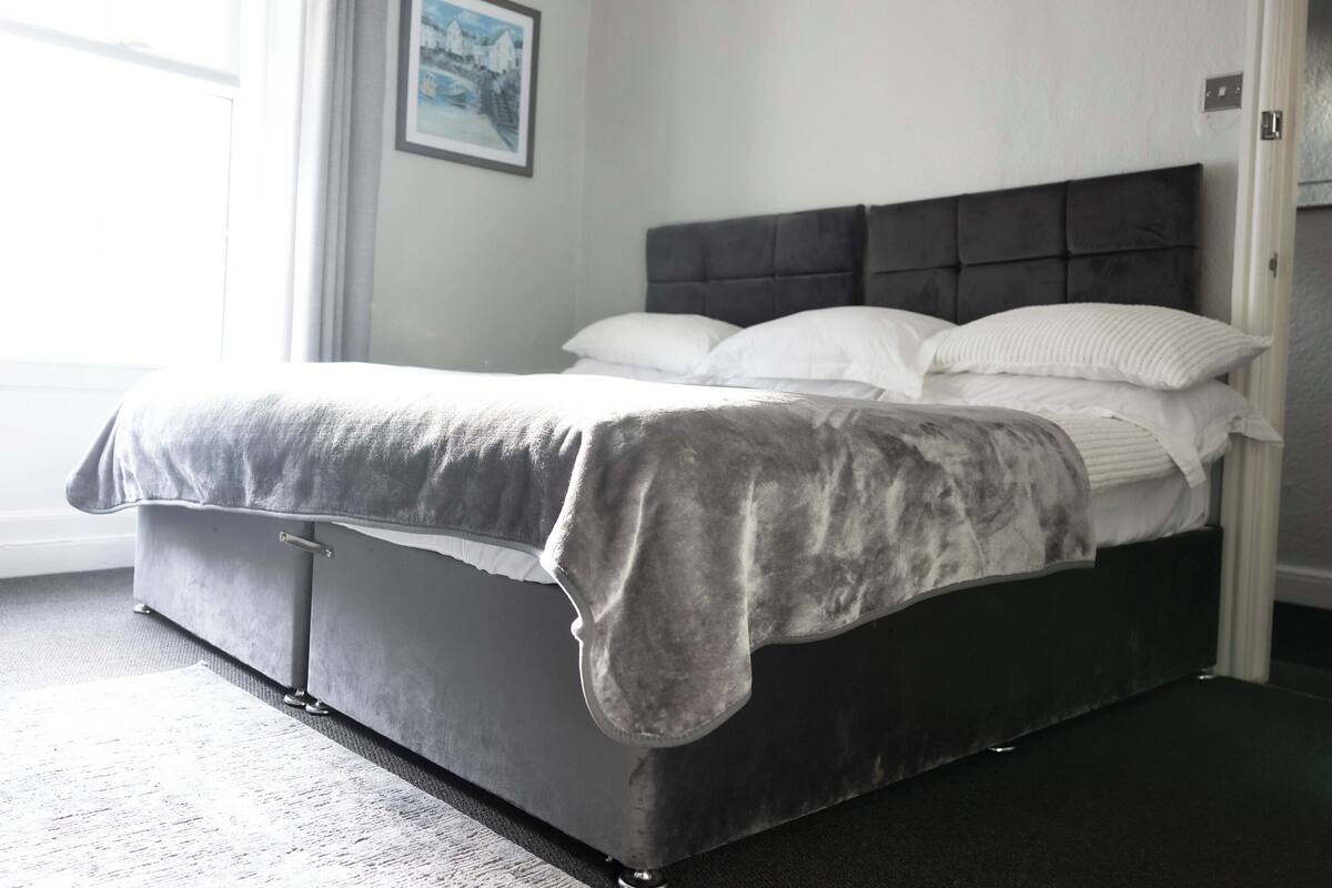 Una camera da letto moderna con un letto grande, una testiera grigia rivestita in tessuto e biancheria da letto bianca.
