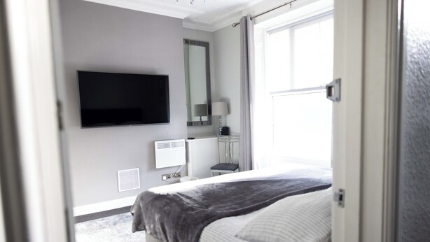 A modern hotel room with a bed, a flat-screen TV, a bedside table, a lamp, and a window with curtains.